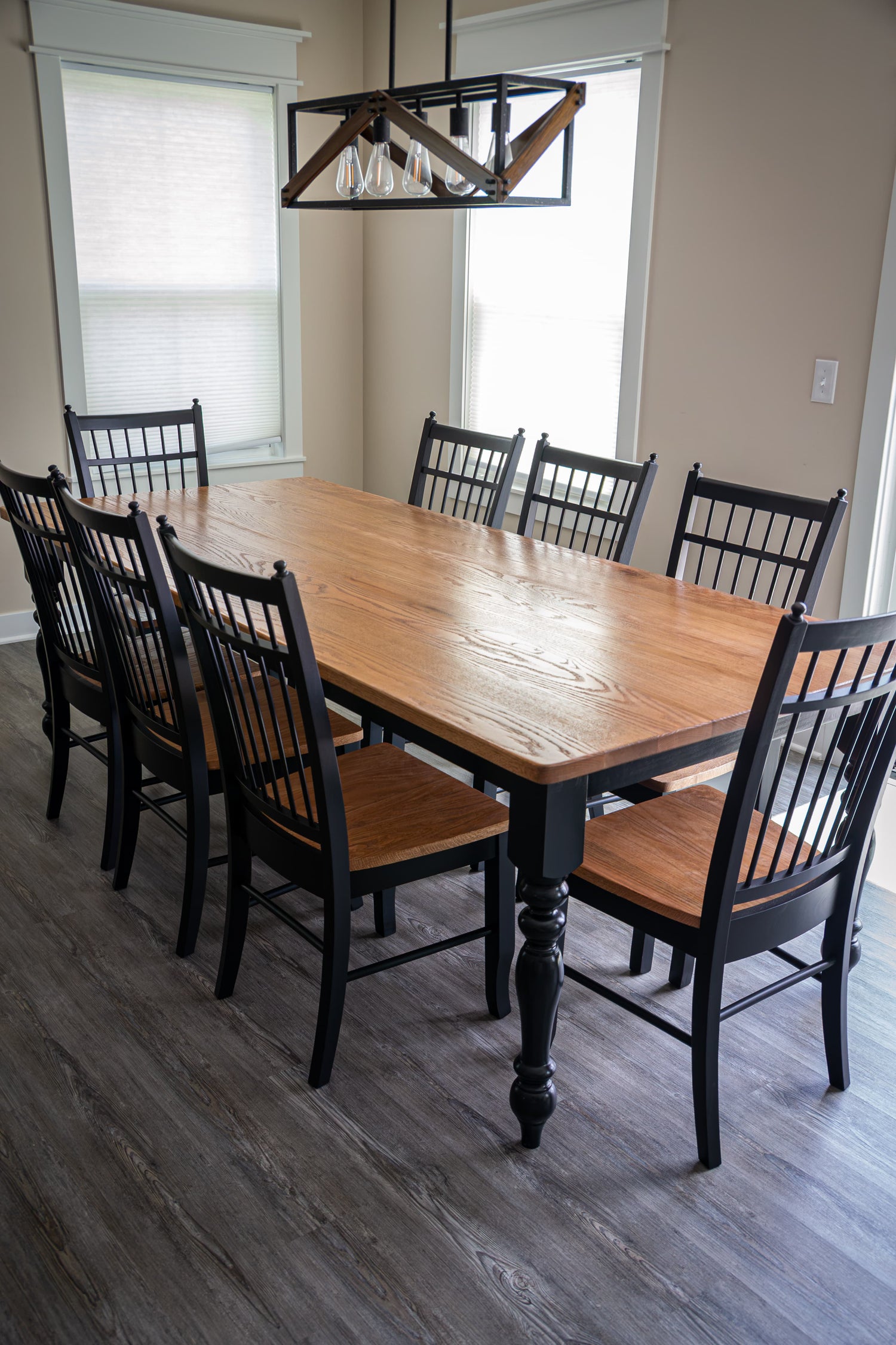 A rectangular wooden dining table with a warm medium-brown wood finish and black spindle-back chairs with brown upholstered seats, shown in a dining room with gray wood-look flooring and a hanging geometric pendant light.