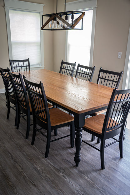 A rectangular wooden dining table with a warm medium-brown wood finish and black spindle-back chairs with brown upholstered seats, shown in a dining room with gray wood-look flooring and a hanging geometric pendant light.