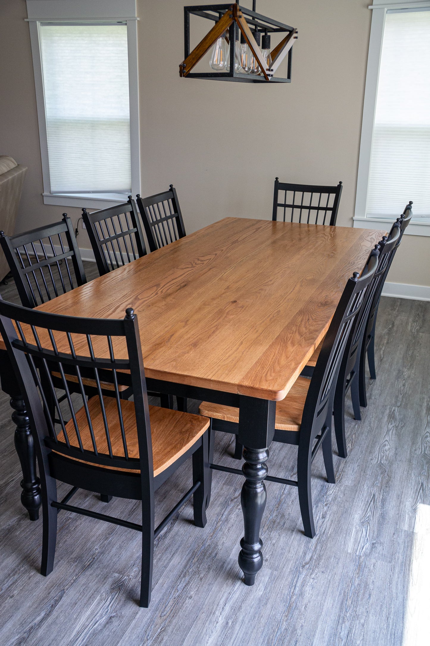 A rectangular wooden dining table with a warm medium-brown wood grain finish, surrounded by black spindle-back chairs with light wood seats, set in a dining room with gray wood-look flooring.