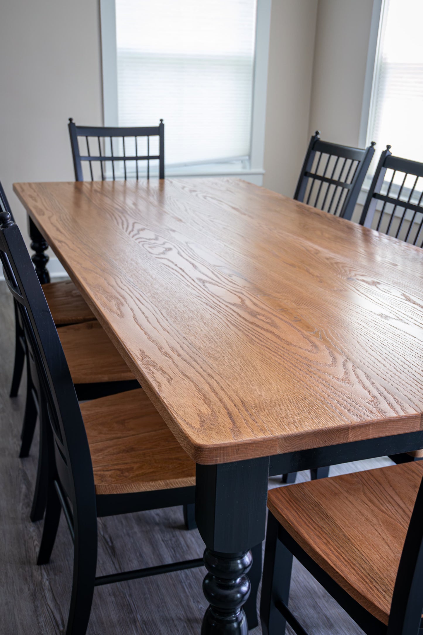 A rectangular wooden dining table with a warm medium-brown oak finish, surrounded by black chairs with wood seats in a bright dining room.