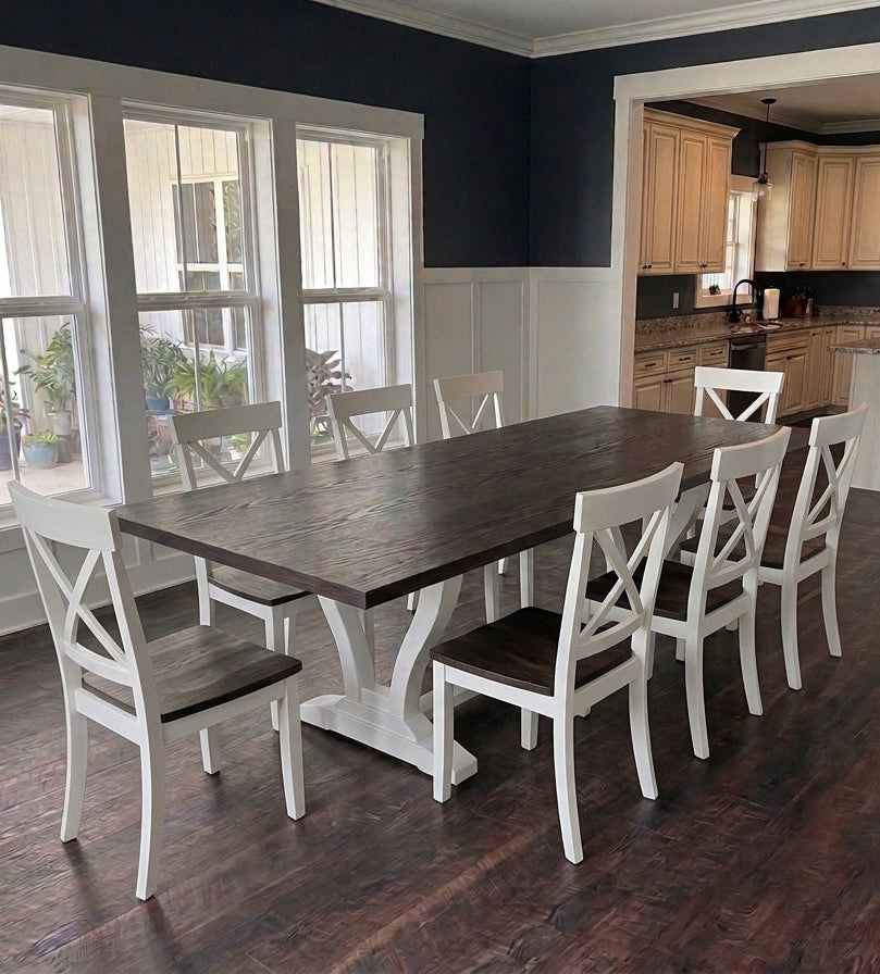A rustic dark wood dining table with six white wooden chairs featuring cross-back designs, set in a dining room with dark wood floors and navy blue walls.